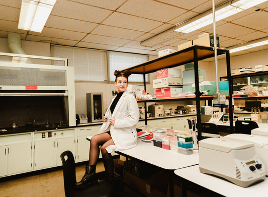 A woman in a lab coat sitting on a bench top in a laboratory surrounded by lab equipment.